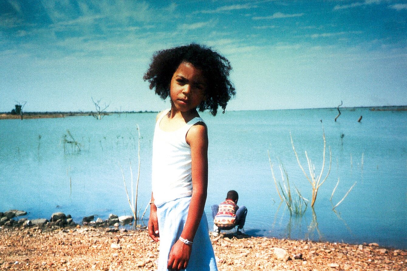 A black girl stands in front of a lake and looks into the camera. Behind her, a boy is sitting on the shore. The sun is shining, the sky and the lake are blue. 