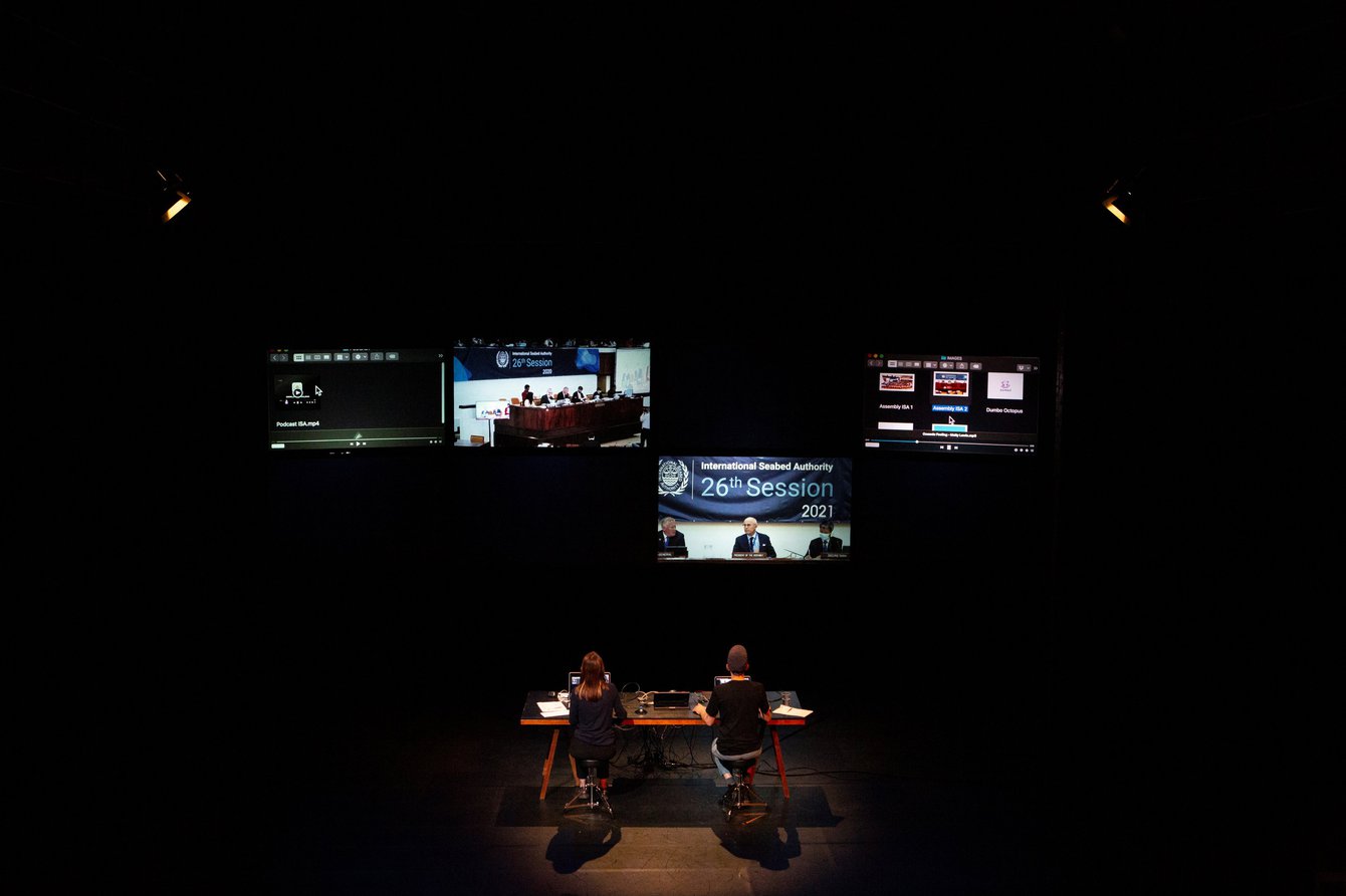 Verschiedene Bildschirme vor schwarzer Wand. Zwei Personen sitzen mit dem Rücken zum Publikum am Schreibtisch vor Laptops.