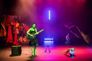 There is a person on stage. She is wearing green color on her entire body. She is playing the guitar and sings into a microphone. In the background there is someone riding on rollerblades. This person is pulling a large, shiny cape behind them.