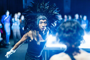 A person with a wig of protruding feathers stands on a stage and is bathed in bluish light while looking an audience member in the eye.