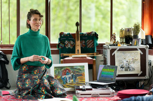 A performer squats in front of objects that she will soon be using in her artistic work: A painting, a pontic lyre and a laptop with music software. 