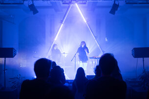 Zwei Musikerinnen stehen auf einer Bühne. Der Raum ist in blaues Licht und Nebel gehüllt. Scheinwerfer und Leuchtröhren werfen dramatisches Licht auf die Bühne.