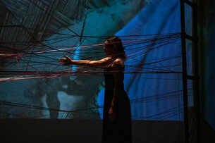 Priyakshi Agarwal on stage between taut yarn. The light is shallow, barely lighting up the set and the performer.