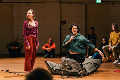 Zwei Performer:innen befinden sich zwischen dem Publikum. Ein:e Performer:in steht in der linken Bildhälfte und blickt in den Raum. Der:die andere Performer:in kniet auf einem Sitzsack. Vor dem:der Performer:in steht ein Ventilator. Der Wind zerrt an den Kleidern und den Haaren des:der Performer:in. Der:die Performer:in hält sich ein Mikrofon an den Mund und blickt angestrengt in Richtung der Kamera.