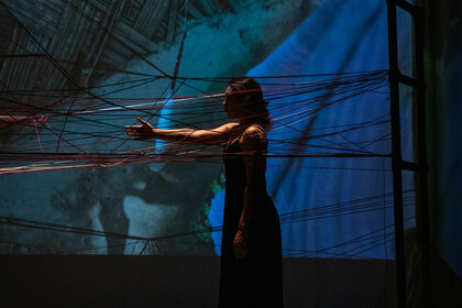 Priyakshi Agarwal on stage between taut yarn. The light is shallow, barely lighting up the set and the performer.