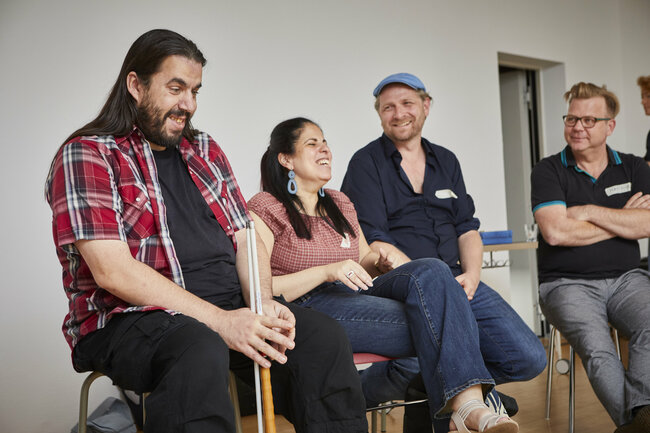 Auf dem Foto sitzen vier Menschen nebeneinander und lachen oder lächeln, während sie miteinander sprechen. Sie wirken entspannt und gesellig. Alle sitzen auf Stühlen in einem hellen Raum.