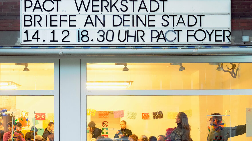 Das Bild zeigt die Außenansicht eines Veranstaltungsortes mit großen Glasfenstern. Oben an der Fassade ist ein Schild mit der Aufschrift „PACT WERKSTADT BRIEFE AN DEINE STADT 14.12 | 18.30 UHR PACT FOYER“ angebracht. Durch die Fenster ist eine Veranstaltung zu sehen, bei der Menschen an Tischen sitzen, sich unterhalten und anscheinend gemeinsam arbeiten oder basteln. Der Raum ist hell beleuchtet, mit bunten Dekorationen an den Wänden, darunter farbige Papiermotive.