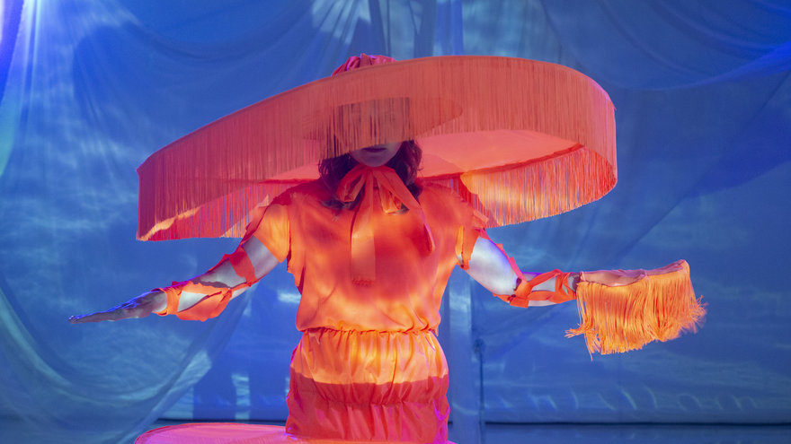 Eine Figur mit Kostüm in Orange und rieseigem runden Hut, der an einen Lampenschirm erinnert vor blauem Hintergrund.
