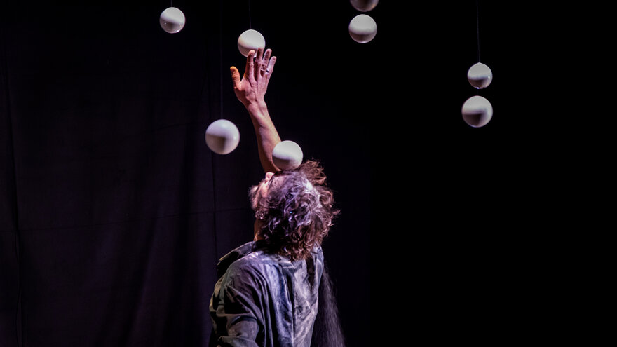 Jongleur Tobias Piero Dohm performt vor einem dunklen Hintergrund. Er streckt den Arm in die Höhe. Um ihn herum schweben mehrere Helle kugelartige Objekte.