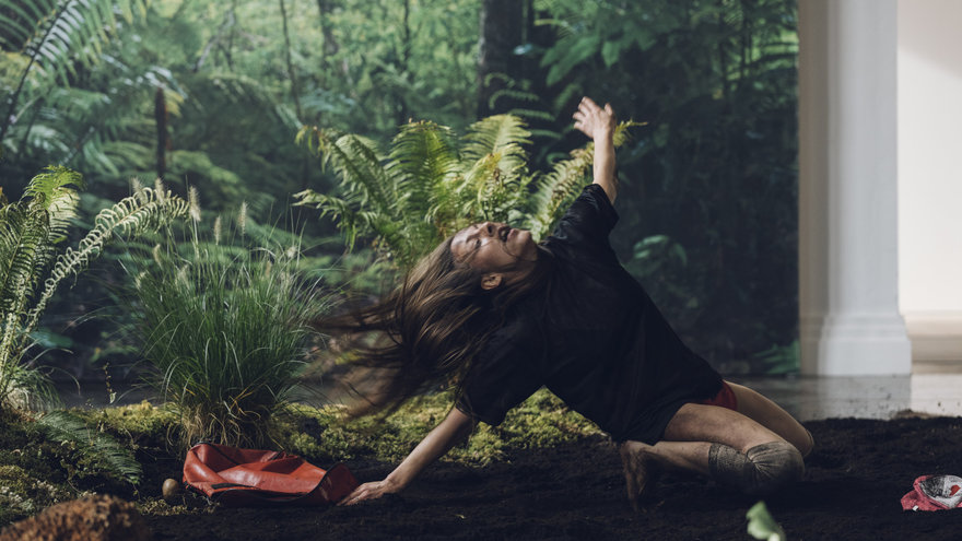 Eine Performerin inmitten einer einer Anordnung aus Erde, Pflanzen, Steinen, wirft sitzende ihre Haare zurück, ein Arm ausgestreckt.