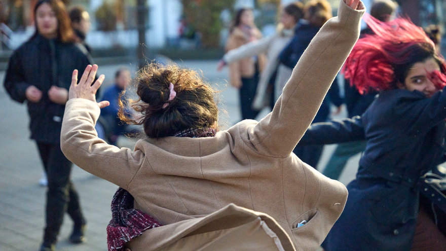 Person von hinten in Bewegung mit ausgestreckten Armen, andere tanzende Personen auf einem Marktplatz